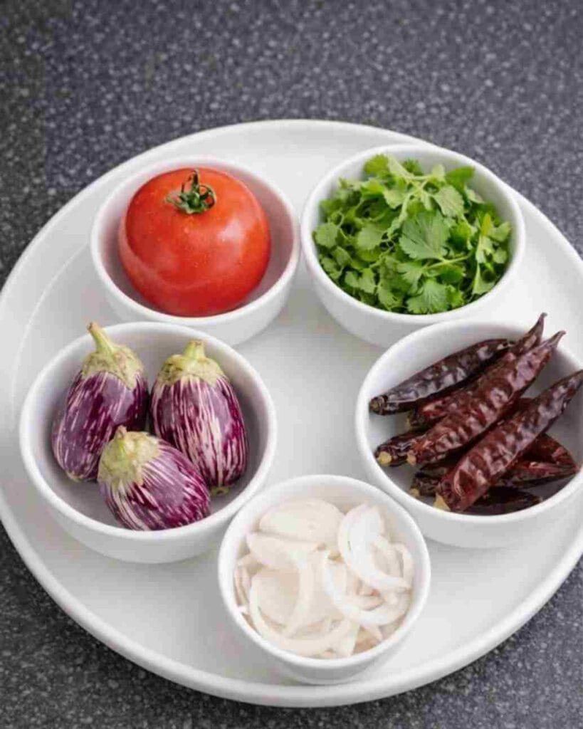 A plate with a tomato, small brinjals, dry red chilies, sliced onions, and fresh coriander leaves in small bowls.