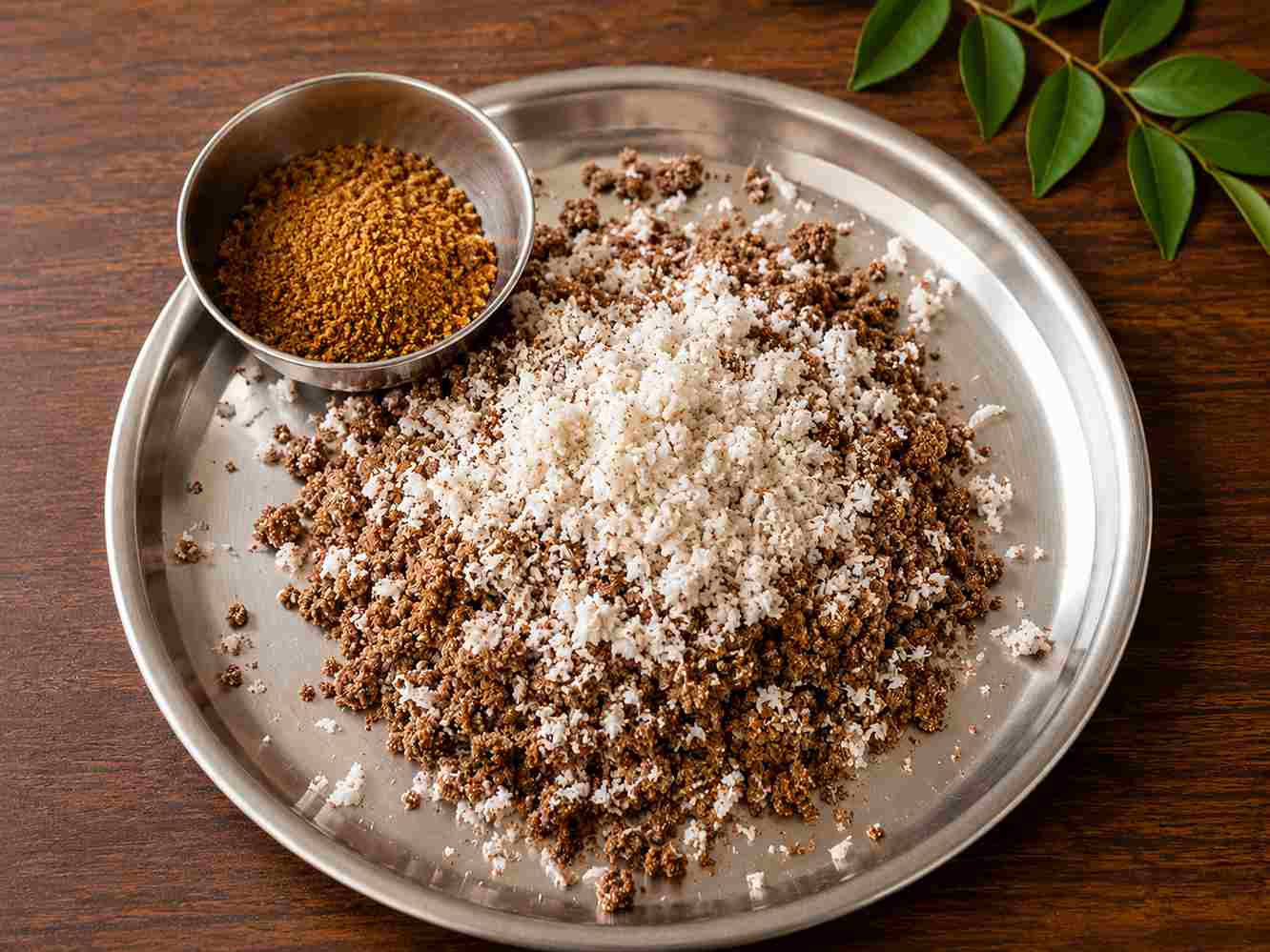 Soft and fluffy ragi puttu recipe served with fresh grated coconut and nattu sakkarai on a stainless steel plate.