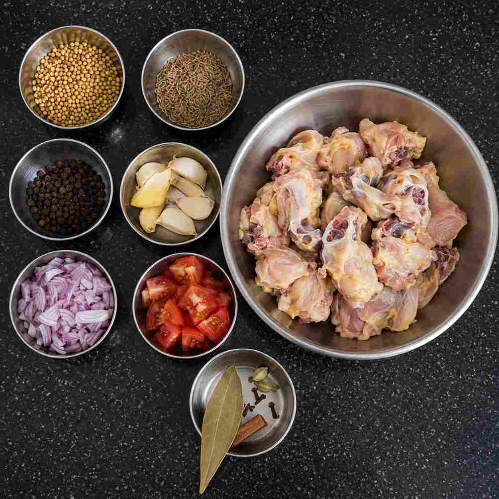 Raw chicken, coriander seeds, cumin, black pepper, ginger, garlic, onion, tomato, and whole spices arranged in bowls.